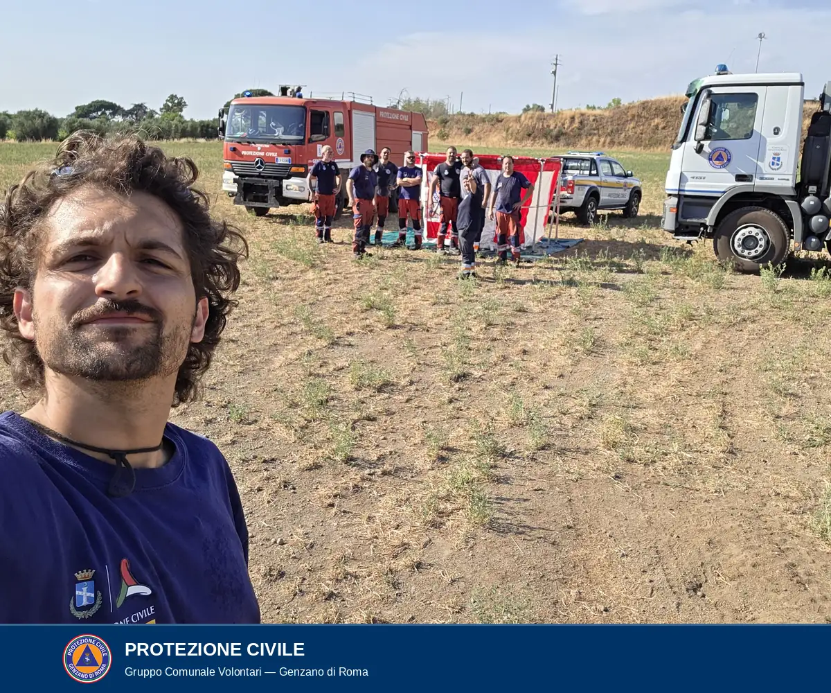 Un volontario in maglietta blu in primo piano; alle sue spalle la squadra schierata accanto alla vasca montata, l'autocarro antincendio rosso, un pick-up bianco e l'autobotte bianca.