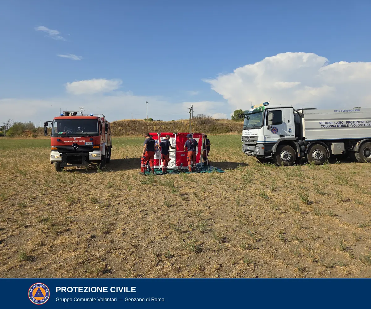 Campo di stoppie con, a sinistra, un autocarro antincendio rosso con la scritta Protezione Civile Città di Genzano di Roma; al centro alcuni volontari montano una vasca in telo rosso e bianco su un telo di base verde; a destra un autocarro bianco con la scritta Regione Lazio Protezione Civile Colonna Mobile Volontariato.