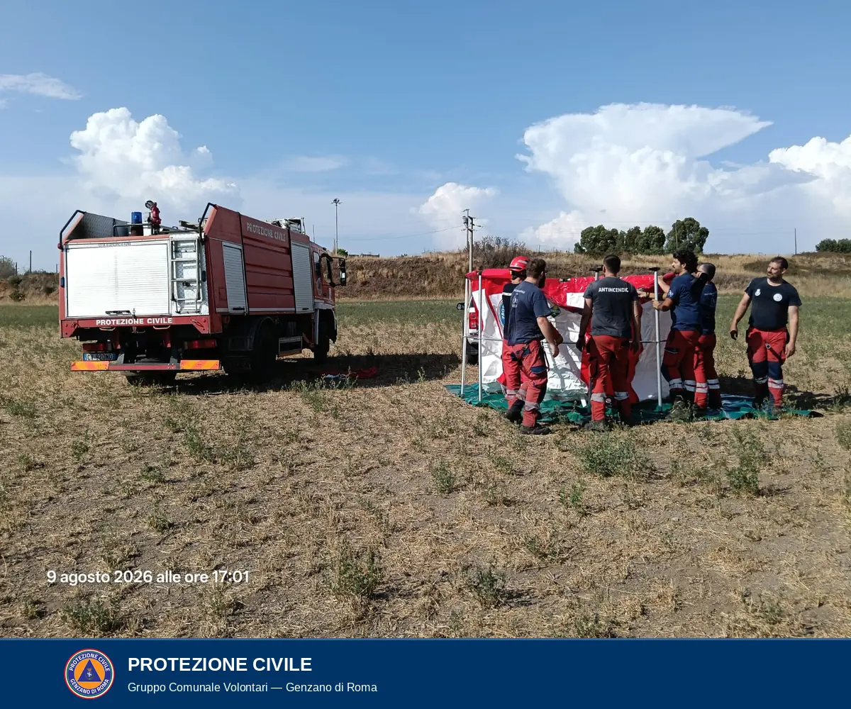 Il telaio metallico della vasca montato in verticale con il telo agganciato ai montanti; alcuni volontari lo sostengono; a sinistra l'autocarro antincendio rosso visto da dietro.