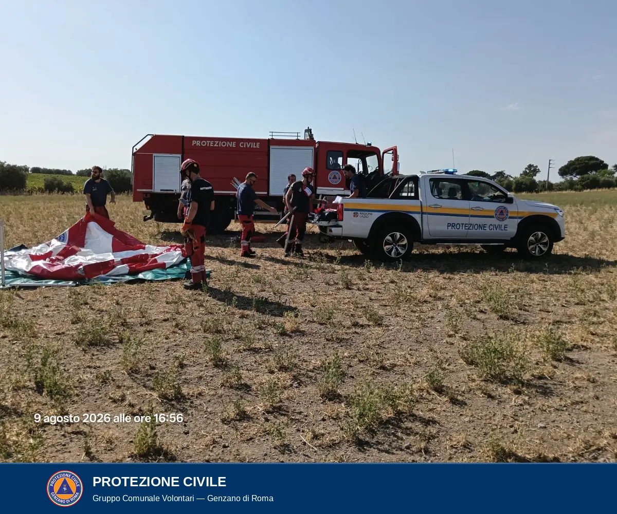 Volontari sistemano il telo rosso e bianco della vasca steso su un telo di base verde, in un campo; sullo sfondo l'autocarro antincendio rosso e un pick-up bianco della Protezione Civile.