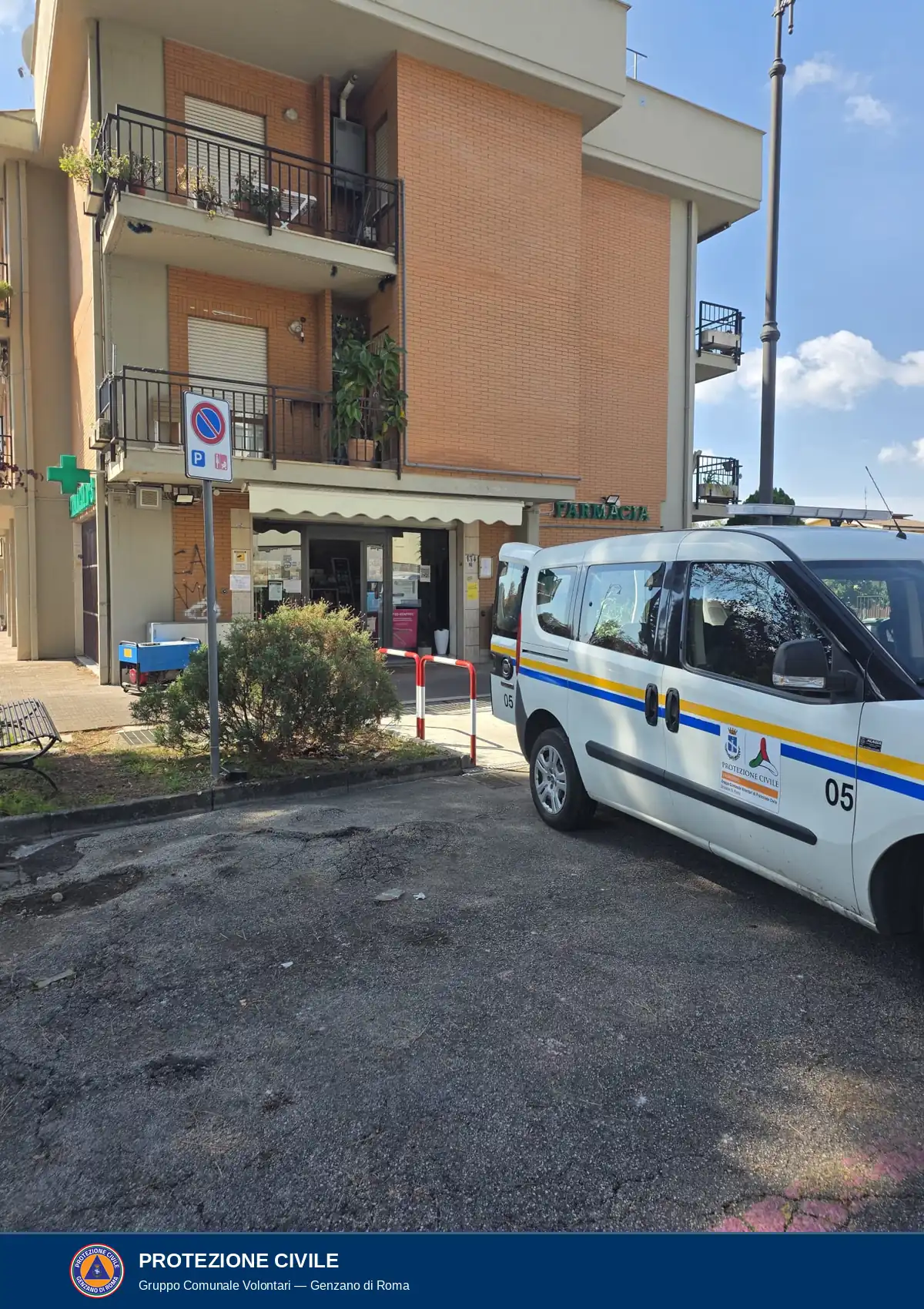 Ingresso di una farmacia sotto un palazzo in mattoni, con una tenda bianca e la scritta Farmacia sulla destra. In primo piano è parcheggiato un Fiat Doblò bianco della Protezione civile con il numero 05 e le strisce gialle e blu. Sul marciapiede, a sinistra di un cespuglio vicino all'ingresso, si vede un generatore blu.