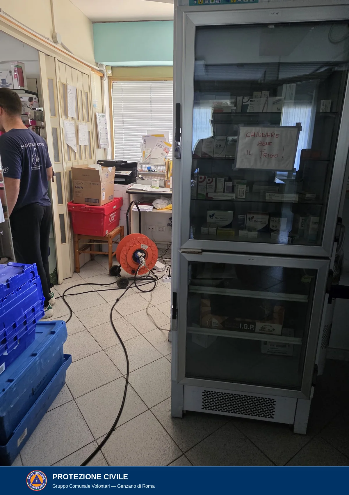 Interno della farmacia: un frigorifero con le ante di vetro e gli scaffali con confezioni di farmaci, con un cartello scritto a mano Chiudere bene il frigo. Sul pavimento un cavo nero arriva da un avvolgicavo arancione. A sinistra, visto di lato, un volontario con la maglietta blu scura con la scritta Protezione civile.