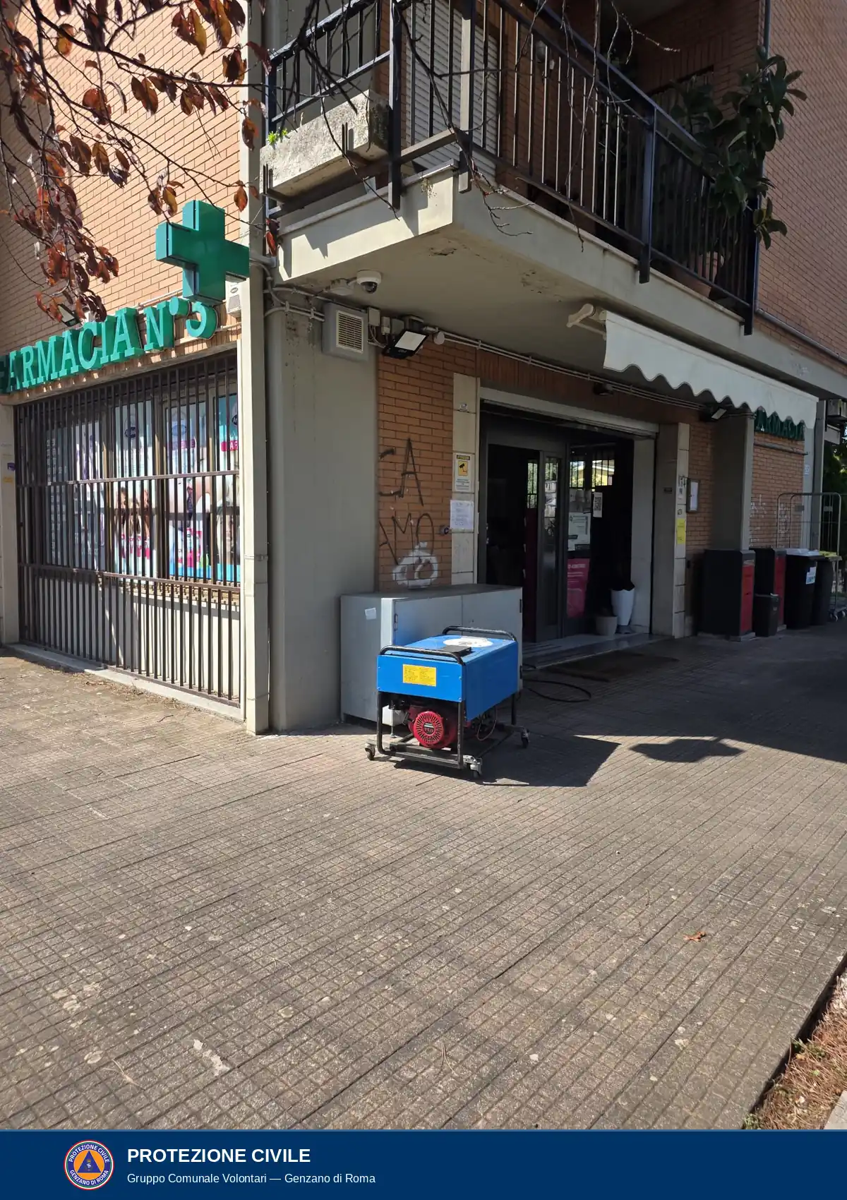 Generatore con carter blu, motore rosso e telaio nero su ruote, sul marciapiede accanto all'ingresso della farmacia. Un cavo nero va verso la porta. A sinistra l'insegna verde Farmacia n. 3 con la croce verde.