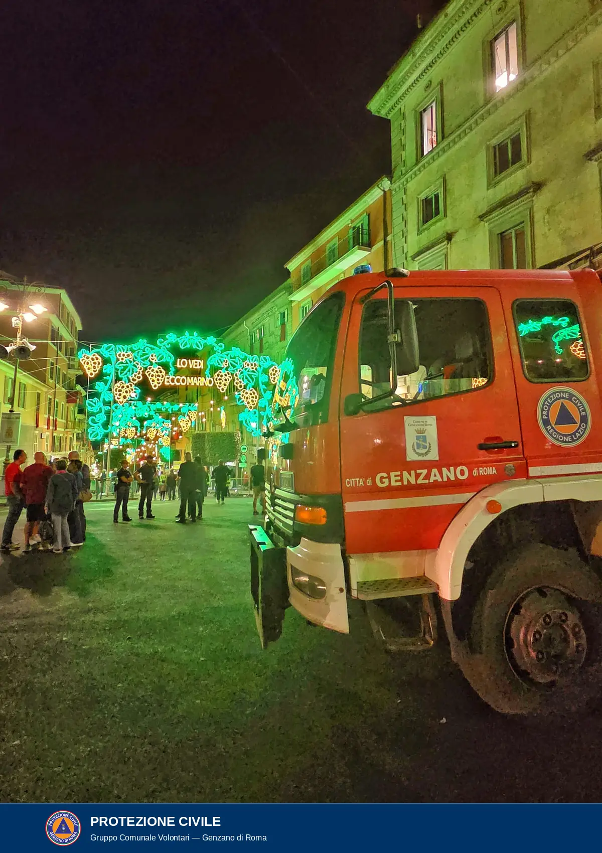Di sera, la cabina e la parte anteriore di un autocarro rosso con la scritta Città di Genzano di Roma, lo stemma del Comune e il logo Protezione Civile Regione Lazio, fermo in una strada tra i palazzi. Più avanti, luminarie verdi con grappoli d'uva dorati e la scritta Lo vedi ecco Marino; sullo sfondo alcune persone in piedi.