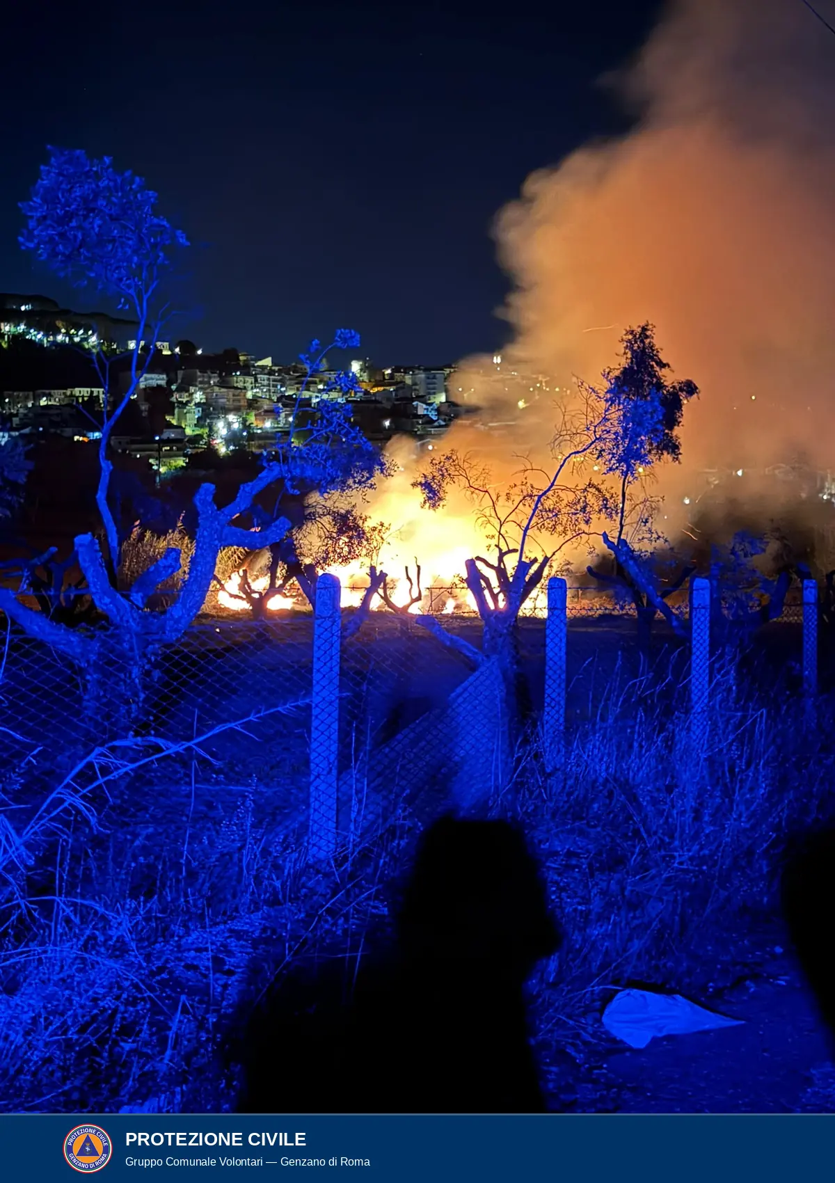 Di notte, oltre una recinzione a rete con paletti di cemento, fiamme alte e fumo arancione fra alberi e sterpi; sullo sfondo le case illuminate di un centro abitato; in primo piano erba e alberi in luce blu e due sagome scure.