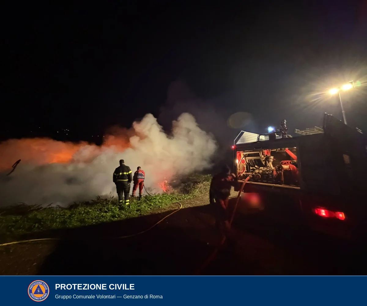 Di notte un vigile del fuoco e un volontario in divisa rossa e blu, di spalle, dirigono una lancia verso una nube di fumo bianco e arancione; a destra un mezzo antincendio con il vano posteriore aperto e una persona che maneggia la manichetta.