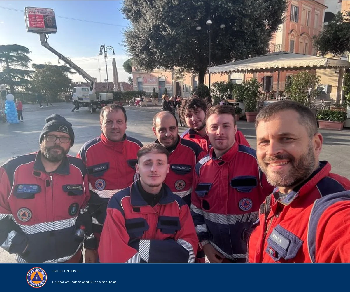 Sette volontari del Gruppo, con le uniformi rosse e blu, posano insieme in piazza Frasconi. Sullo sfondo una piattaforma aerea solleva uno striscione.