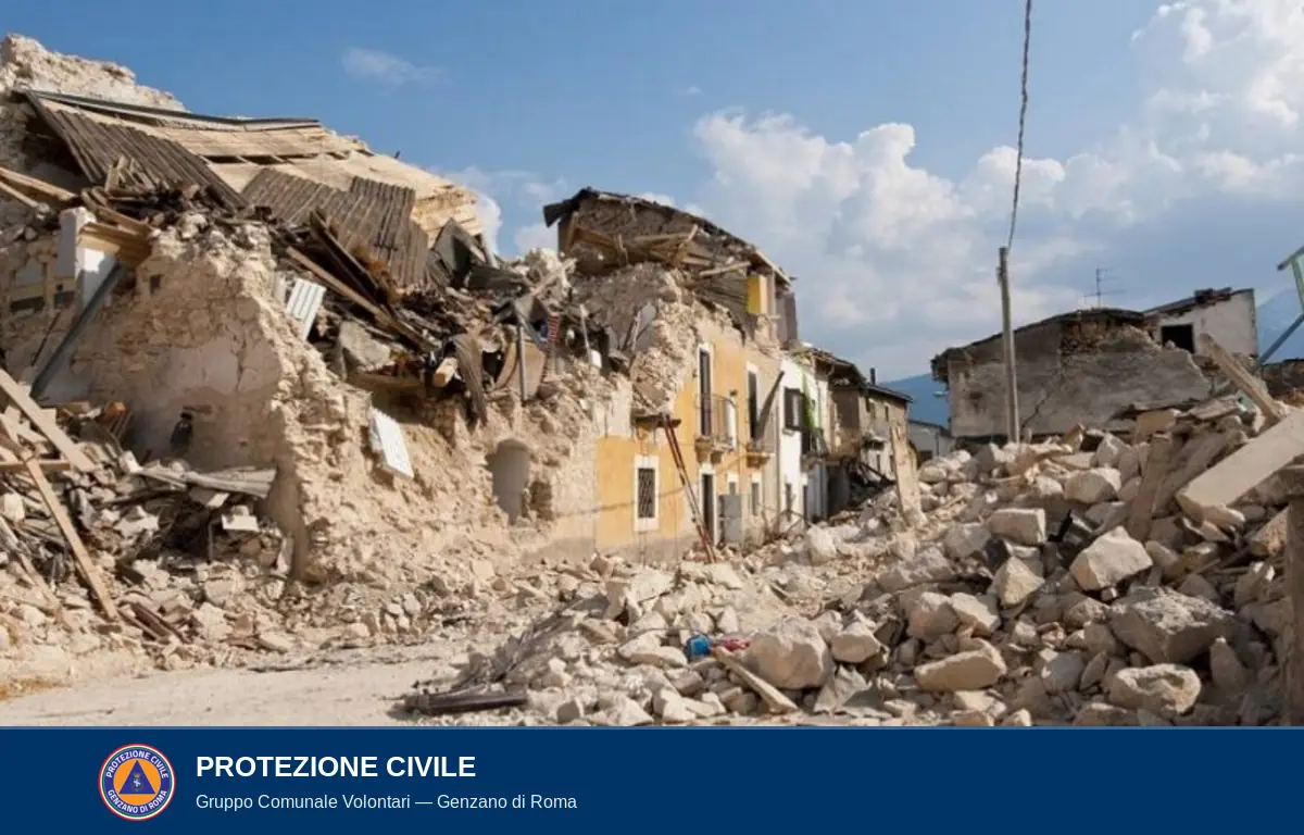 Foto a colori del 2016: vista di una via di un piccolo centro storico marchigiano dell'Appennino dopo la sequenza sismica del Centro Italia. A sinistra una grande casa in pietra è completamente sventrata: il tetto è crollato, le travi di legno spezzate sporgono dalle pareti rimaste, l'intonaco è caduto. Sul centro della via, cumuli di macerie — pietre, calcinacci, tegole — formano una distesa irregolare. A destra altri edifici lesionati con crepe profonde. Sopra, cielo azzurro estivo.