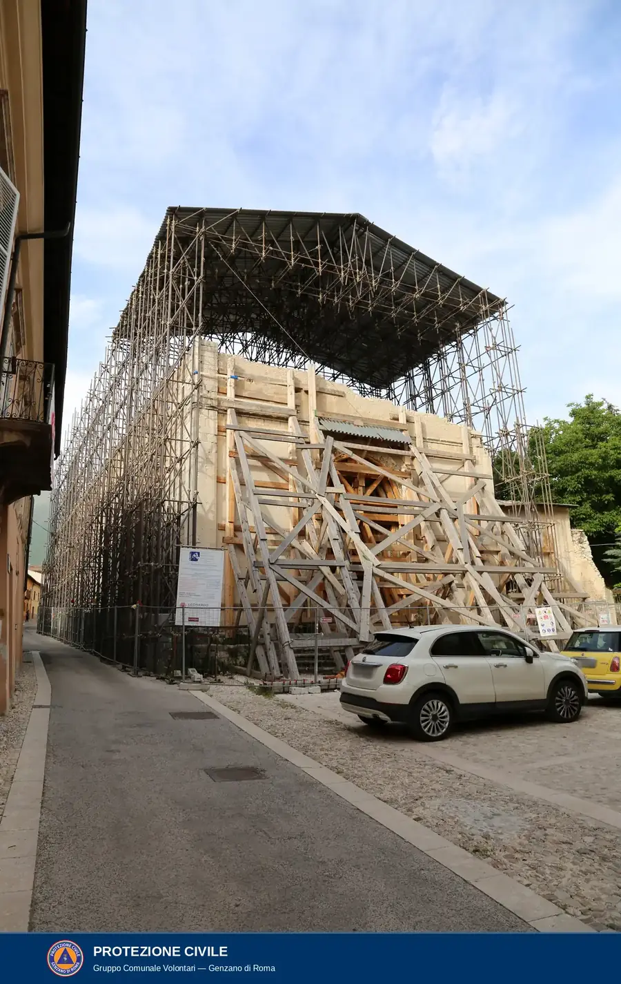 Rudere della chiesa di Sant'Agostino a Norcia dopo il terremoto: le mura sopravvissute sono puntellate da un'imponente struttura di tavole di legno incrociate e sovrastate da un'ampia tettoia di protezione su ponteggi metallici; auto parcheggiate sulla strada in primo piano